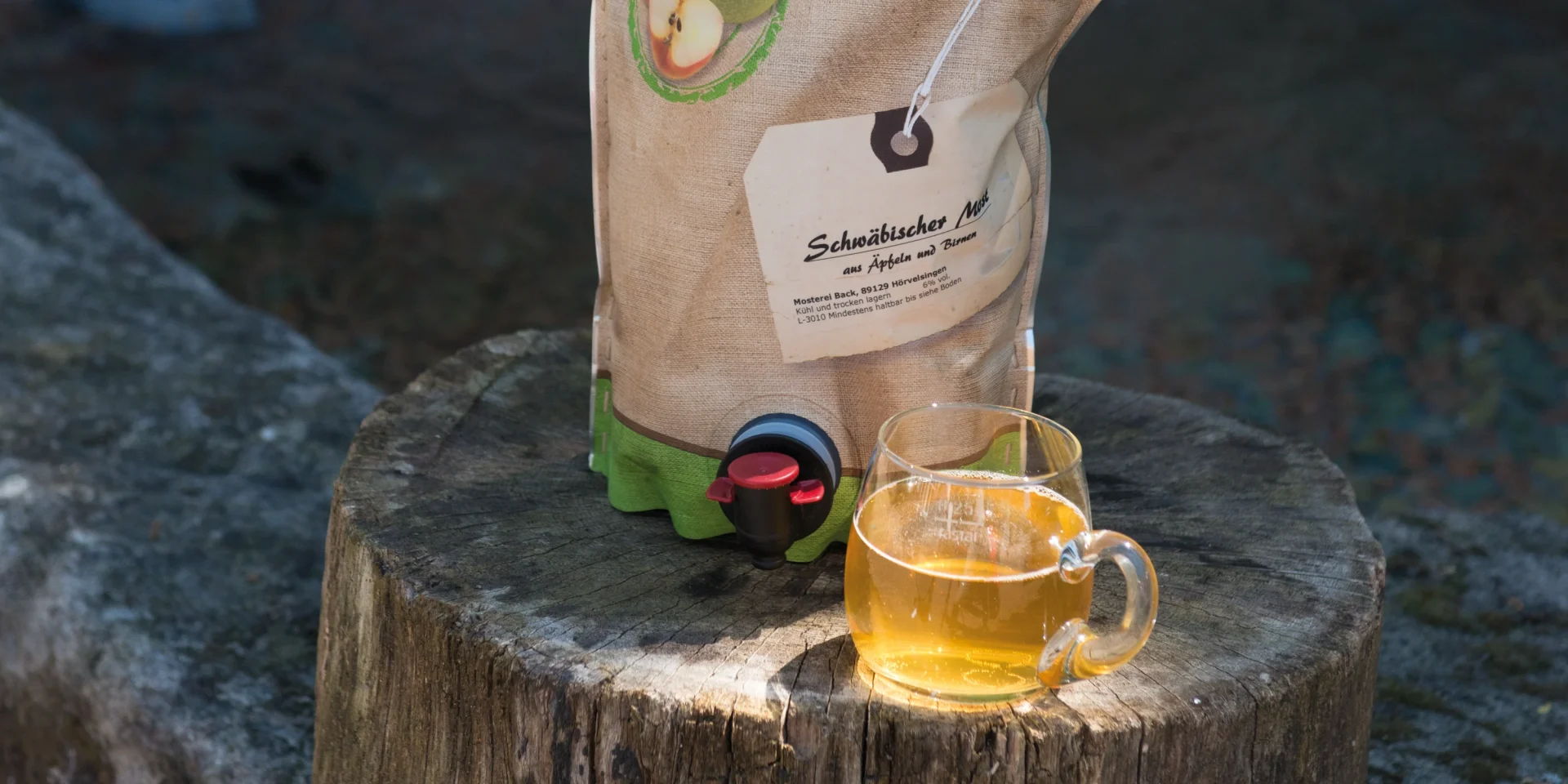 Auf einem Baumstumpf steht ein Glas "Schwäbischer Most" vor der Verpackung mit Zapfhahn.