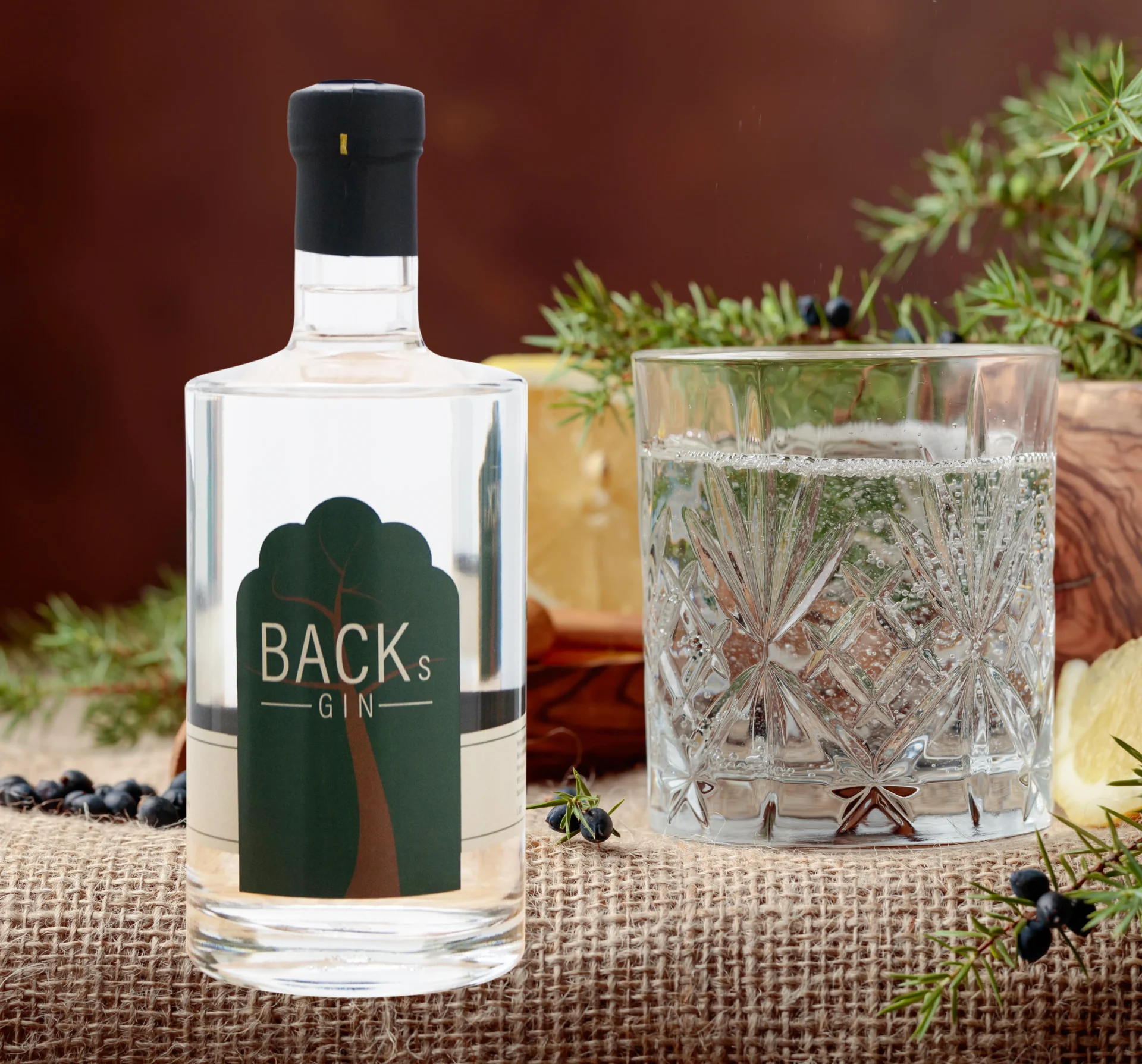 Eine Flasche Backs Gin steht auf einem Tisch, daneben ist ein volles Glas und im Hintergrund ist eine Zitrone und Wacholder
